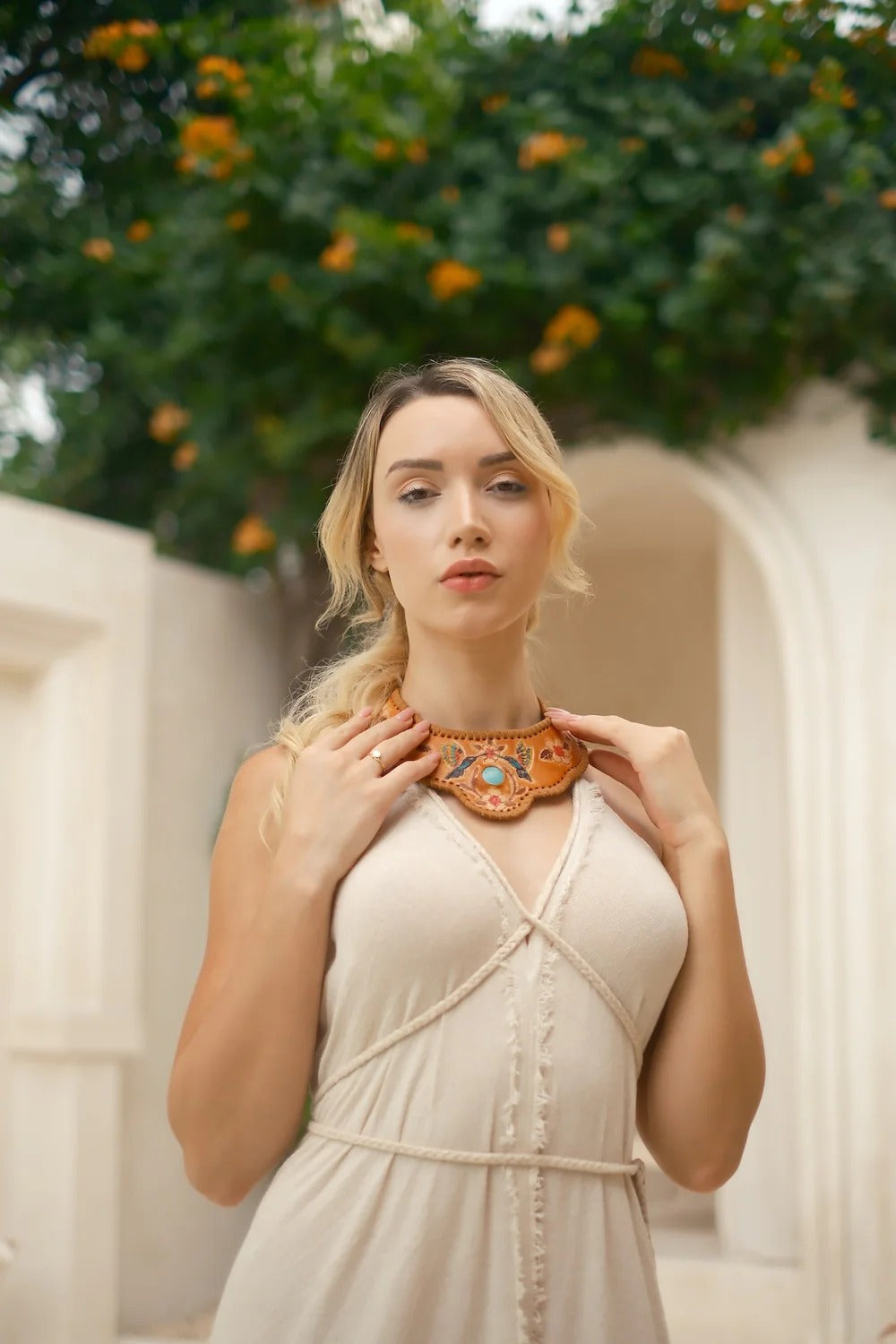 Woman wearing a beige dress and brown necklace in an outdoor setting with greenery.