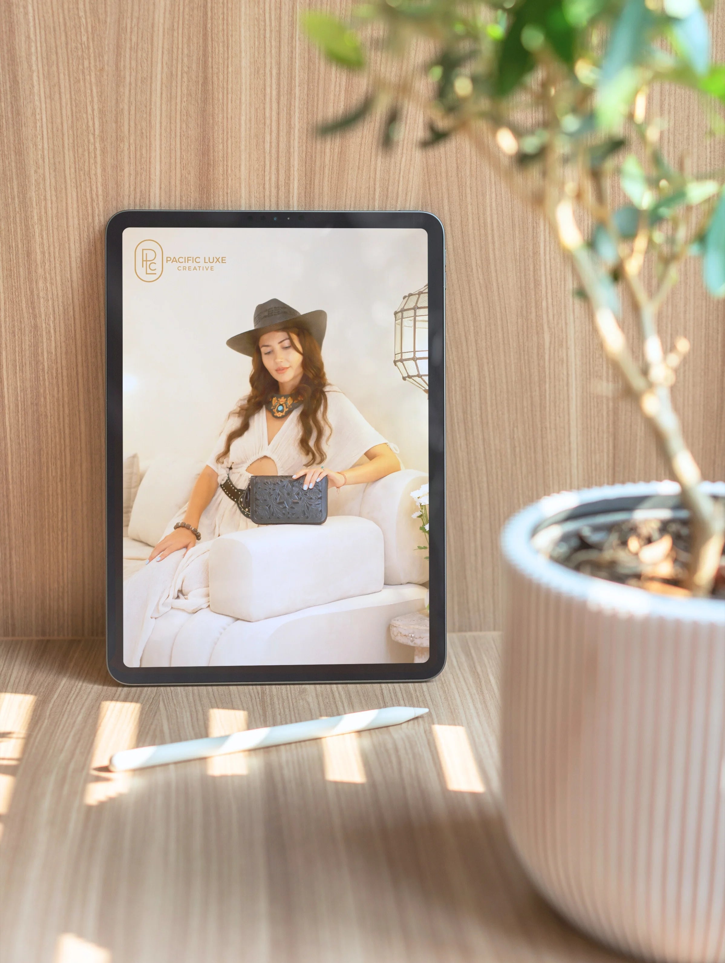 Tablet showcasing Pacific Luxe Creative brand photography of a woman in a hat and dress, styled on a wooden surface with soft greenery glow in the background.