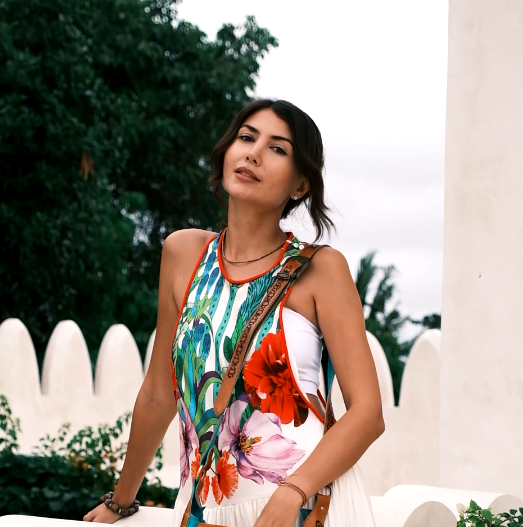 Person in vibrant floral dress posing outdoors against scalloped white wall with tropical trees